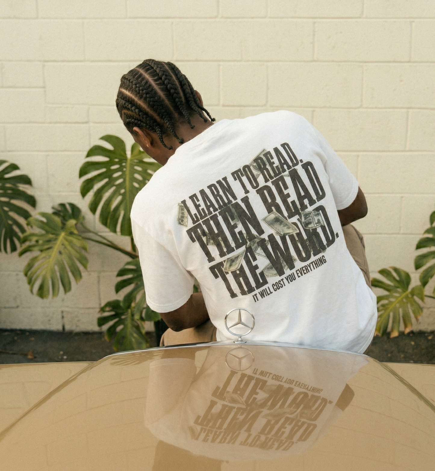 Person wearing a white t-shirt with text and a logo, sitting on a reflective surface with plants in the background.