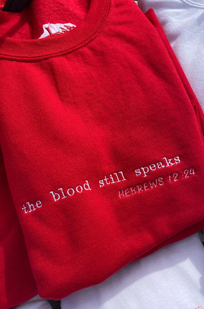 A red sweatshirt with the text 'the blood still speaks' and the Bible verse 'Hebrews 12:24' embroidered on it.