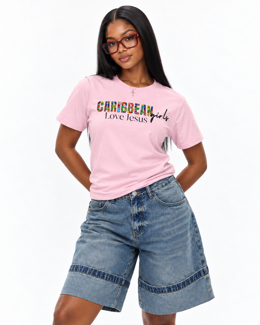 A woman modeling a light pink graphic t-shirt with 'CARIBBEAN girls Love Jesus' printed on the front, paired with denim shorts against a plain white background.