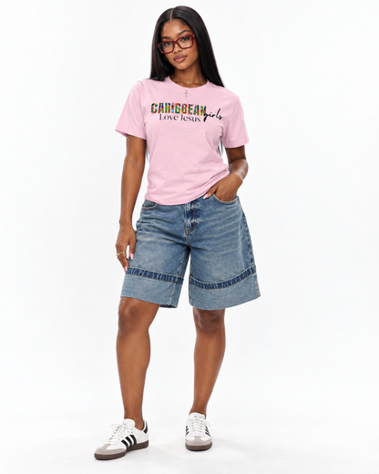 A woman modeling a light pink t-shirt with 'CARIBBEAN girls LOVE jesus' printed across the chest, paired with denim shorts and white sneakers, against a plain white background.