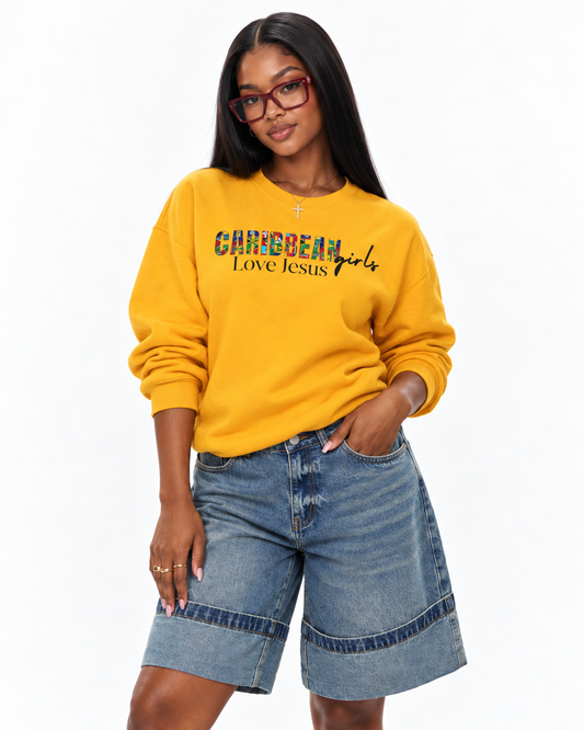 A woman modeling a mustard yellow sweatshirt with 'caribbean girls Love Jesus' printed across the chest in a multicolor floral pattern, paired with denim shorts, against a plain white background.