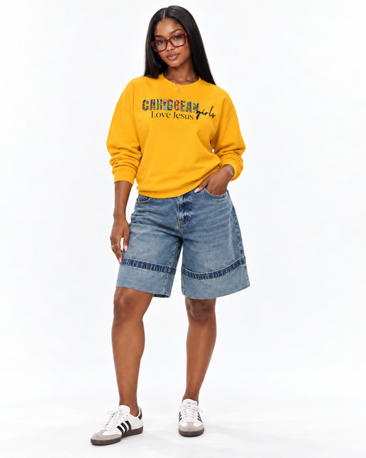 A woman modeling a bright yellow sweatshirt with 'caribbean girls love jesus' printed on the front, paired with light-wash denim shorts and white sneakers, against a plain white studio background.